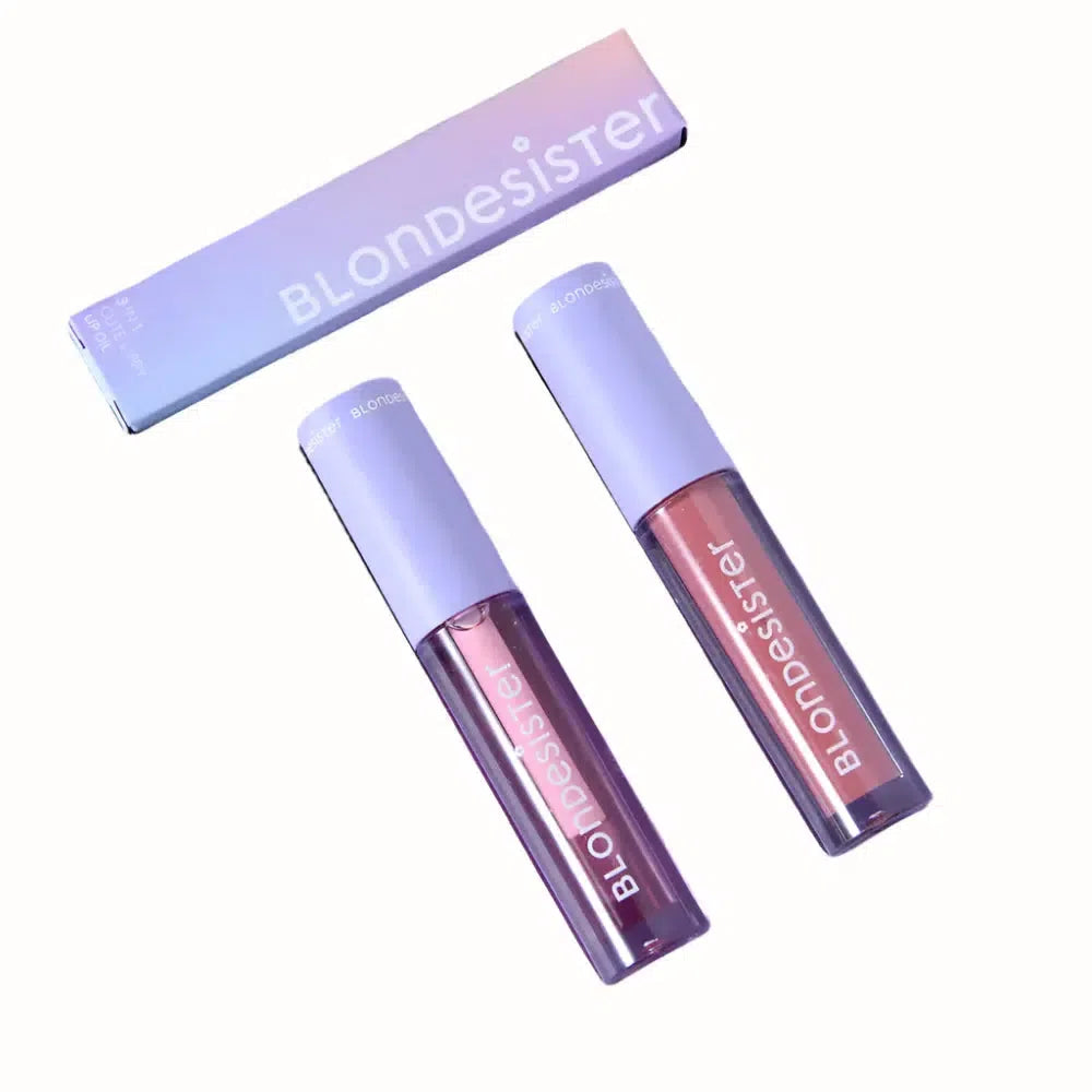 A sleek set of two BlondDesister lip glosses nestled in a purple box with a soft pastel gradient lid. Each gloss is housed in a clear tube with a lavender cap, labeled with the BlondDesister logo in white. The set is designed to offer a luxurious, vibrant color with a high-shine finish.