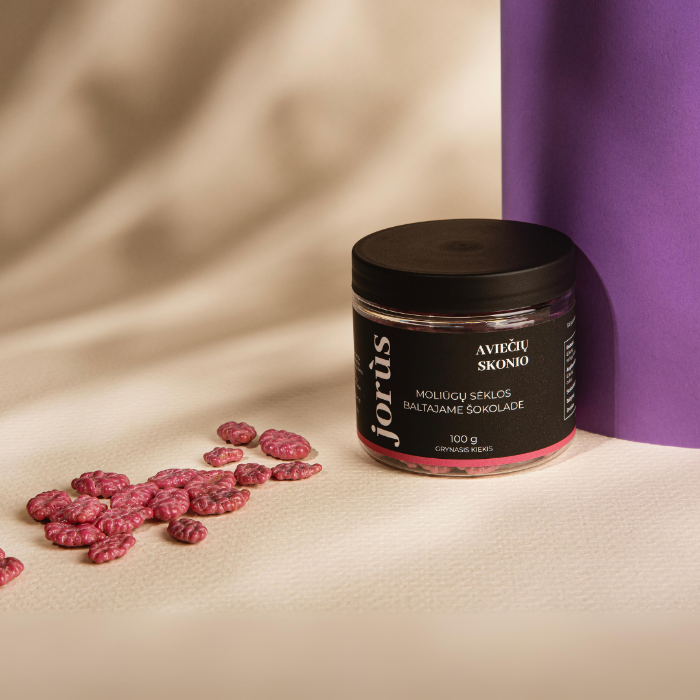 A jar of Jorus raspberry-flavored seeds coated in white chocolate. The packaging features the flavor in Lithuanian "Aviečių Skonio" and includes details such as the product weight (100g) and the brand name, with the jar showcasing the pink-colored seeds.