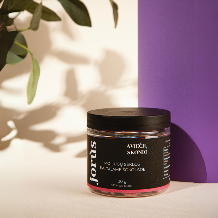 A jar of Jorus raspberry-flavored seeds coated in white chocolate. The packaging features the flavor in Lithuanian "Aviečių Skonio" and includes details such as the product weight (100g) and the brand name, with the jar showcasing the pink-colored seeds.
