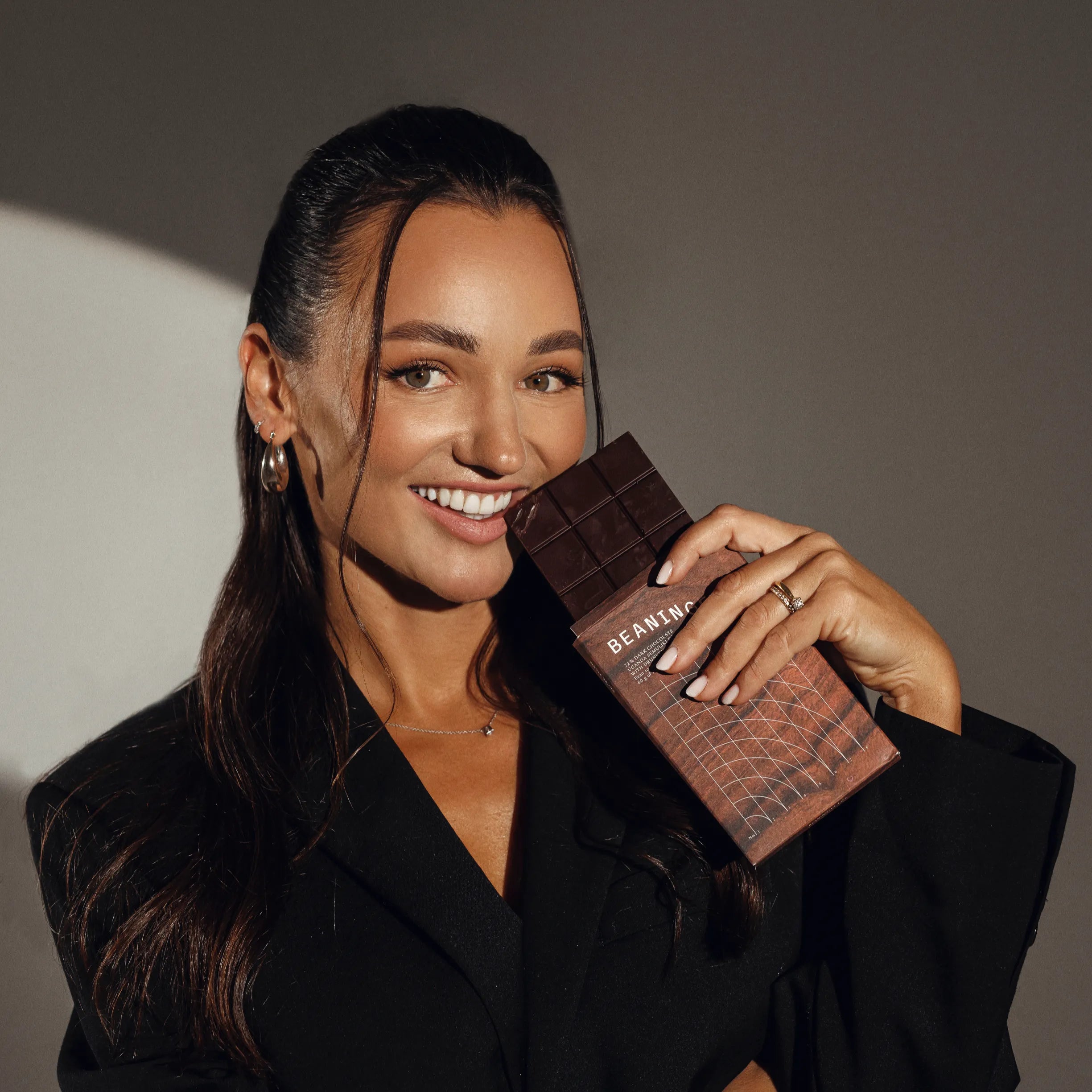 A woman in a black blazer smiling while holding a chocolate bar. She poses confidently, showing her sparkling smile and wearing elegant jewelry, with a natural and relaxed vibe.